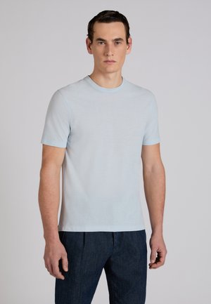 Mężczyzna stojący przodem, ubrany w prostą, jasnoniebieską koszulkę z krótkim rękawem i ciemne spodnie na neutralnym tle.