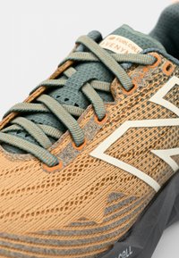 Scarpa da trail running con tomaia in mesh di colore tan e grigio, lacci teal, suola testurizzata e distintivi accenti arancioni. Presenta un grande logo.