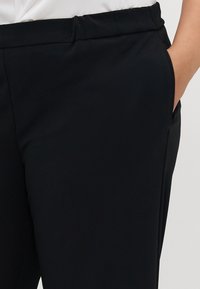 Person wearing black tailored pants with hand in right pocket, paired with a tucked-in white top, shown from waist to mid-thigh.