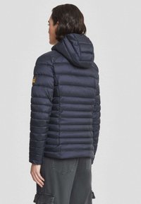 Persoon in een marineblauwe gewatteerde hoodie jas met een logo patch op de linkerarm, gecombineerd met donkere cargobroek, gezien van achteren.