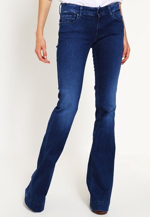 Dunkelblaue Slim-Fit-Jeans mit einem leichten Schlag am Beinende, getragen von einer Person, die vor einem einfarbigen hellen Hintergrund steht.