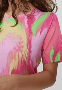 Femme portant un t-shirt en coton à manches courtes avec un motif abstrait rose, vert et jaune, avec des cheveux bruns bouclés lâches partiellement visibles.