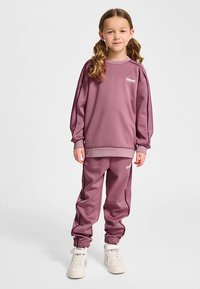 Sweat-shirt de sport violet et jogging assorti en tissu doux, avec des poignets côtelés et un logo sur la poitrine et la cuisse. Baskets blanches.