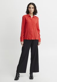 Camicia rossa a maniche lunghe con bottoni e taschino sul petto, abbinata a pantaloni neri a gamba larga e stivali alla caviglia, su uno sfondo neutro.