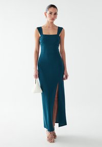 Blågrön, ribbad midi-klänning med bred fyrkantig urringning, figursydd överdel och lårhög slits i sidan; kombinerad med en vit triangelväska.