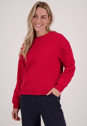 Rode sweatshirt met een ronde hals, ribbels aan de mouwen en onderaan. Heeft een klein geborduurd logo op de borst. Gecombineerd met een donkere broek.