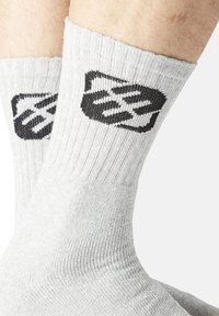 Gros plan sur des jambes portant des chaussettes côtelées gris clair avec un logo géométrique noir près des mollets sur fond uni.