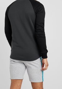 Maglietta nera a maniche lunghe con corpo grigio, polsini a coste, abbinata a pantaloni corti grigi chiari con strisce blu ai lati, texture liscia.