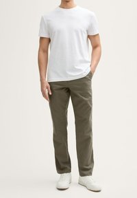 Homme debout, main droite dans la poche, portant un t-shirt blanc uni, un pantalon vert olive et des baskets blanches sur un fond clair.