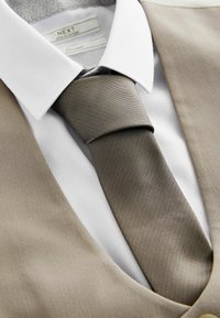 Veste beige avec une texture lisse, chemise blanche à col avec un motif en losanges et une cravate taupe, présentant un design diagonal épuré.