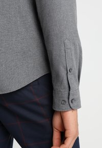Camicia grigia con bottoni, realizzata in un tessuto leggermente strutturato, caratterizzata da un singolo bottone ai polsi e un orlo curvo, abbinata a pantaloni scuri con motivo.