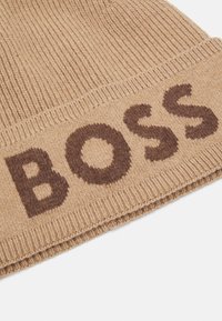 Beige pletená čepice s ohnutým lemem a tmavě hnědým textem "BOSS". Texturovaný žebrovaný design, měkký materiál a čisté okraje.