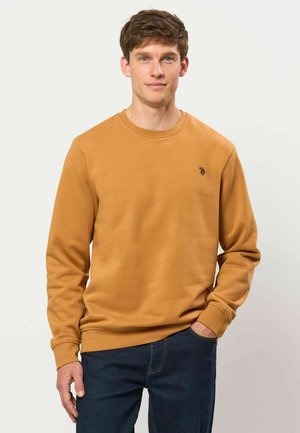 Man met een lichte huid en kort bruin haar draagt een mosterdkleurig shirt met lange mouwen en donkerblauwe spijkerbroek, staat met één hand in de zak.