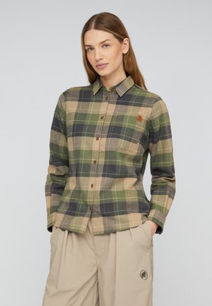 Femme portant une chemise à carreaux verts et beige boutonnée avec un pantalon beige, debout devant un fond neutre, les mains dans les poches.