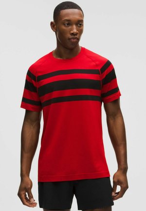 Hombre con camisa roja de manga corta con tres rayas horizontales negras en el pecho y las mangas, combinada con pantalones cortos negros.