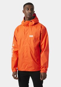 Oranje waterdichte jas met capuchon, voorzien van elastische cuffs en een voorzak. Witte logo op de borst en mouw. Glad textuur.