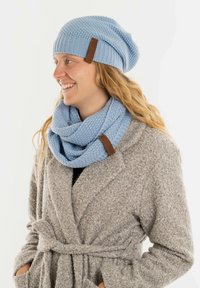 Lichtblauwe gebreide beanie en oneindige sjaal met een gestructureerd patroon. De sjaal is om de nek gewikkeld. Bruine leren accenten zijn zichtbaar.
