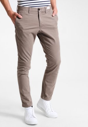 Mand iført beige slim fit bukser og hvide høje sneakers, hænderne i lommerne, foran en ensfarvet lys baggrund.