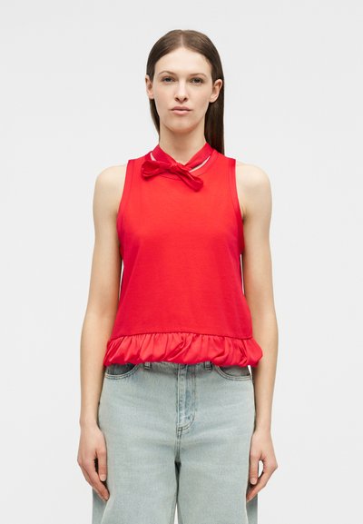 Γυναίκα που φοράει αμάνικη κόκκινη μπλούζα με φιόγκο κοντά στον λαιμό και βολάν στο τελείωμα, συνδυασμένη με ανοιχτόχρωμο τζιν, στέκεται μπροστά σε απλό φόντο.