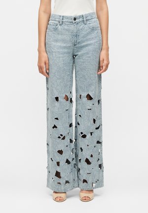 Jasnoniebieskie szerokie dżinsy z kwiatowym haftem z wycięciami, noszone z beżowymi sandałami na obcasie z odkrytymi palcami i białym topem, widok z przodu.