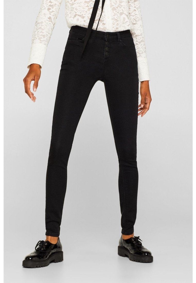 Jeans skinny negros de denim, con una cintura alta, cierre de cinco botones, trabillas para cinturón y un diseño minimalista. Combinados con zapatos negros brillantes.