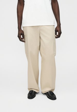 Mand iført beige cargobukser med vidde ben og lynlåslommer, hvid skjorte og sorte loafers med kvaster, stående foran en ensfarvet baggrund.