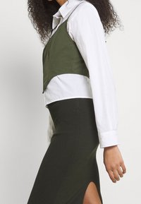 Avklippt, olivgrön väst lagd över en vit skjorta med knappar, i kombination med en lång, figurnära olivgrön kjol med en sidoflik.