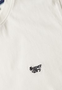 Biała bawełniana koszulka z okrągłym dekoltem, z małym czarnym haftowanym logo "Super Dry" na klatce piersiowej. Gładka faktura.