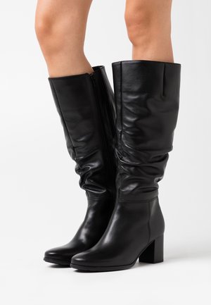 Bottes en cuir noires jusqu'au genou avec des talons bloc portées par une personne, présentant des zips sur le côté et des tiges légèrement froncées.
