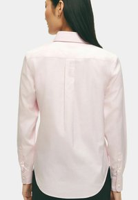 Chemise à manches longues de couleur rose clair avec un col pointu, un détail de pli au dos et des poignets boutonnés. Texture lisse et ourlet arrondi.