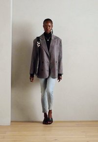 Blazer grigio over-size in tessuto morbido con due grandi bottoni, abbinato a una maglia nera, jeans azzurri chiari e scarpe scivolate nere. Accessori minimi.