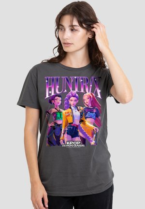 Frau trägt dunkelgraues T-Shirt mit buntem animiertem Trio und dem Schriftzug "HUNTRX K-POP DEMON HUNTERS", richtet ihre Haare, heller Hintergrund.