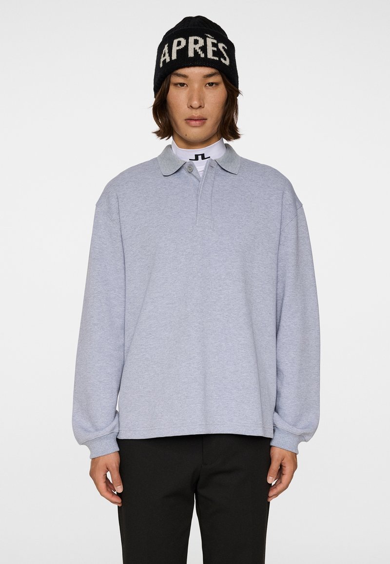 Osoba ubrana w jasnoszarą koszulkę polo z długim rękawem, czarne spodnie oraz czarną czapkę z napisem "APRÈS" w kolorze białym, stojąca na białym tle.