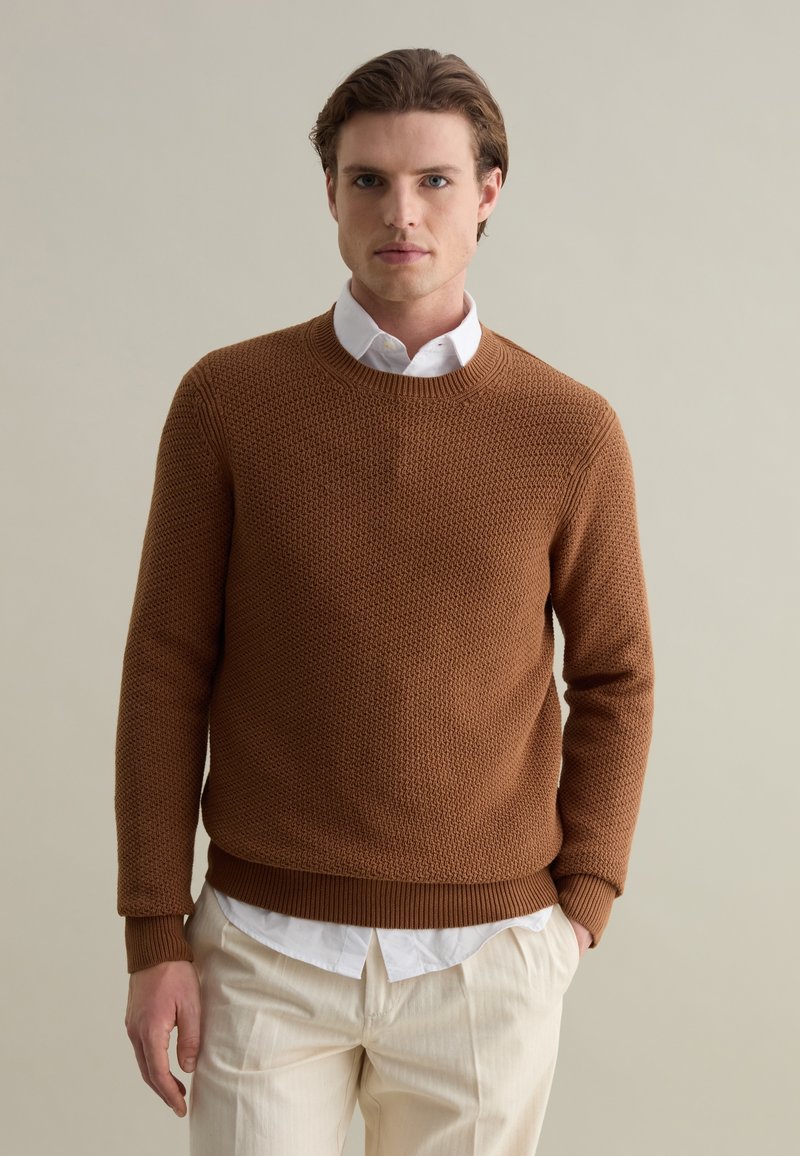 Brown knitted sweater with a textured pattern, round neckline, and ribbed cuffs. Worn over a white shirt with a visible collar.