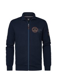 Marinblå zip-up jacka med ett quiltat mönster, kontrasterande blå dragkedja och logotyp patch på bröstet. Har ribbade ärmslut och nederkant.