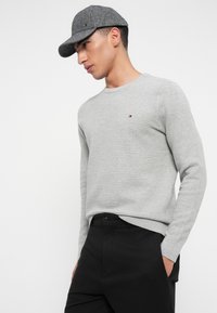 Νεαρός άνδρας που φοράει ένα γκρι υφασμάτινο sweater με στρογγυλή λαιμόκοψη, μαύρο παντελόνι και γκρι καπέλο του μπέιζμπολ, στέκεται με το ένα χέρι στην τσέπη.