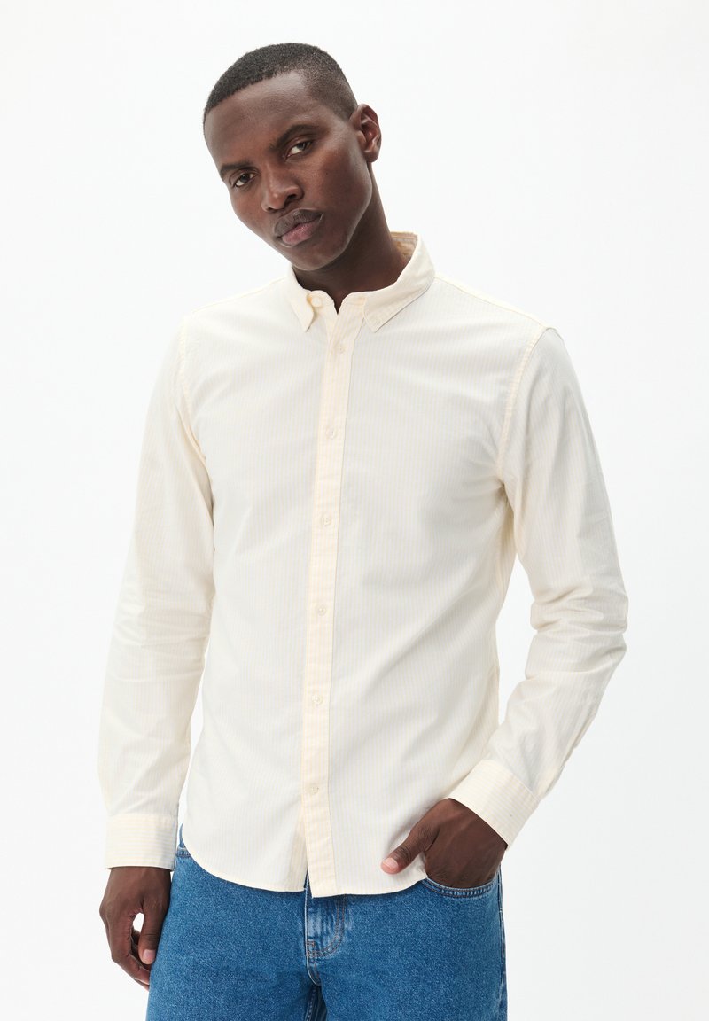 Lysegul skjorte med knaplukning, der har subtile lodrette striber, lange ærmer og krave i klassisk stil, parret med blå jeans.