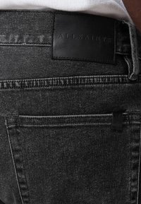 Mustat farkut, joissa on matta nahkainen brändilogo "ALLSAINTS" vyötäröllä sekä huomaamatonta ompelua taskujen lähellä.