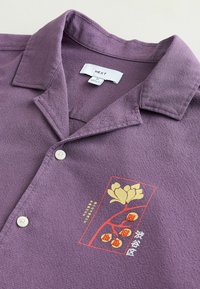 Chemise violette à col avec des boutons blancs et une fleur jaune brodée ainsi que des baies rouges à l'intérieur d'un rectangle rouge sur la poitrine.
