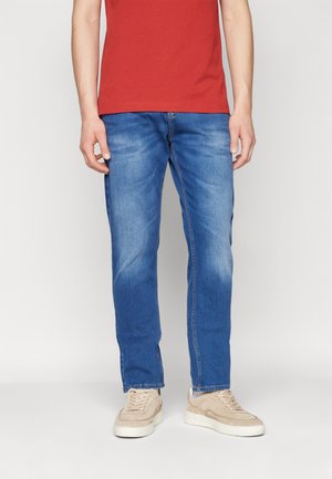 Homme portant un jean bleu, des sneakers beiges et une chemise rouge à manches courtes, debout devant un fond clair uni.