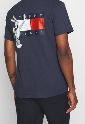 Mężczyzna w granatowej koszulce z postacią Buga Bani trzymającą marchewkę obok czerwono-biało-granatowego logo Tommy Jeans na plecach.