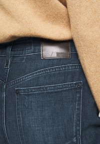 Jeans de mezclilla azul oscuro con una etiqueta de cuero que dice "DRYKORN" y un suéter suave de color marrón claro que cubre parcialmente la cintura.