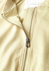 Bomber jacket giallo chiaro con finitura strutturata, dotato di una zip argentata frontale e dettagli a coste sul colletto. Etichetta del marchio visibile all'interno.
