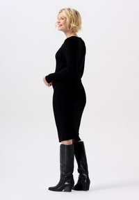 Schwarzes, langärmliges, tailliertes Kleid mit einem mittellangen Saum, kombiniert mit knielangen schwarzen Lederstiefeln, präsentiert vor einem schlichten weißen Hintergrund.