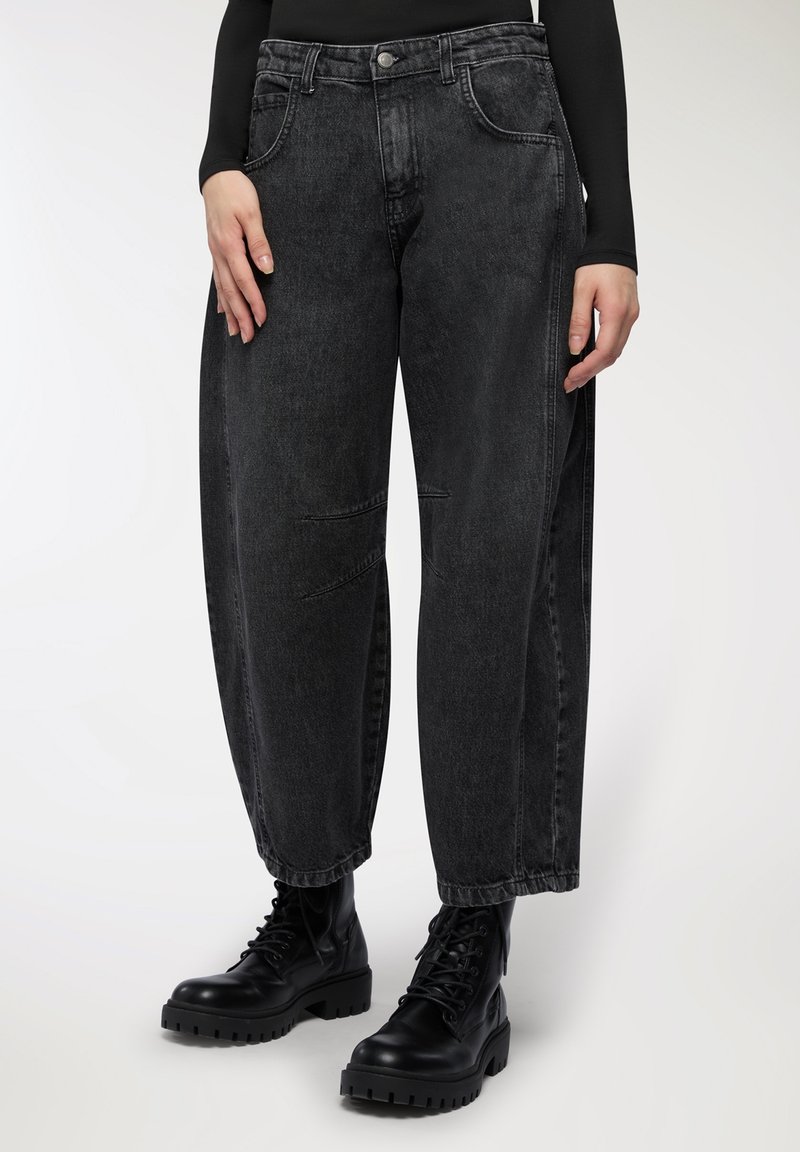 Jeans in denim nero con vestibilità rilassata, dotati di tasche frontali, cuciture in vista e finitura sfumata, abbinati a stivali neri robusti.
