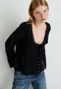 Giovane donna con pelle chiara e capelli rossi, che indossa grandi orecchini a cerchio, una blusa di pizzo nera e jeans blu, in piedi contro uno sfondo bianco uniforme.