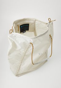 Hvit nylon totebag med glidelås, med gule og svarte snørehåndtak, et romslig interiør og en kontrastetikett inni.