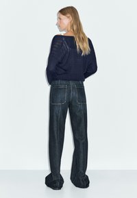 Dunkelblauer gestrickter Pullover mit lockerer Passform, kombiniert mit hoch taillierten, ausgestellten Jeans aus Denim mit sichtbaren Nähten und großen Gesäßtaschen.