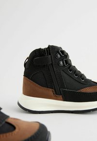Chaussures montantes noires et brunes avec une tige texturée, des lacets, une sangle Velcro et une semelle blanche. Détail de fermeture éclair sur le côté.