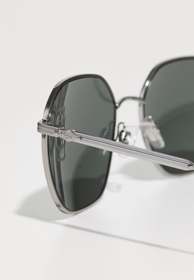 Occhiali da sole con sfumatura grigia, montatura in metallo argento elegante, forma angolare e aste sottili. Punta delle aste trasparente con dettagli del logo.